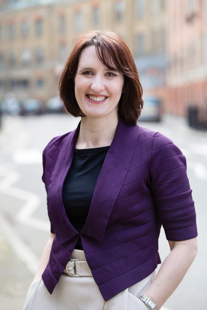 Laura Gibson, a whitenon-disabled woman, standing in the street with her hands in her pockets smiling at the camera